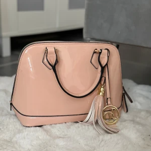 Ljusrosa lackad handväska med tofs - Snygg ljusrosa handväska i lackat material med svarta detaljer och guldfärgade metalldelar. Väskan har två handtag och en dekorativ tofs med guldring och bokstavshänge. Perfekt för dig som gillar en chic och modern stil.