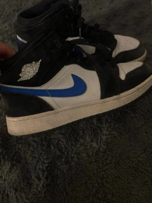 Air Jordan 1 Mid svart/blå/vit - Säljer ett par Air Jordan 1 Mid sneakers i svart, blått och vitt läder. Klassisk Jordan Wings-logga på sidan och blå swoosh. Svart snörning och orange Jumpman-detalj på plösen. Perfekta för dig som gillar streetwear och ikoniska sneakers.