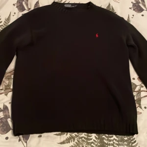 Svart stikad tröja från Polo Ralph Lauren - Säljer en klassisk svart tröja från Polo Ralph Lauren med röd broderad logga på bröstet. Tröjan är tillverkad i 100% bomull och har långa ärmar med ribbade muddar. Perfekt för en stilren look. 