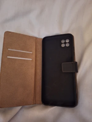 Svart plånboksfodral till mobil - Snyggt svart mobilskal i plånboksmodell med plats för kort på insidan. Utsidan är i konstläder och insidan har en mjukare beige yta. Skalet har magnetlås och öppning för fyrdubbel kamera. Perfekt för dig som vill ha både skydd och funktion i ett. Passar till samsung galaxy a22 5g.