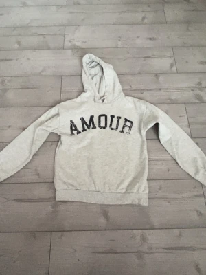 Grå hoodie med AMOUR-tryck - Ljusgrå hoodie med svart texten 'AMOUR' framtill och en svart stjärna på ryggen. Klassisk huva och ribbade muddar vid ärmar och nederkant. Perfekt för en avslappnad och trendig look. Mjuk bomullsblandning för extra komfort. Den är nopprig. 