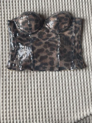 Leopardmönstrad paljett korsettopp - Säljer en snygg leopardmönstrad korsettopp med paljetter. Toppen är axelbandslös och har vadderade kupor för extra stöd. Perfekt för dig som vill sticka ut med ett djurmönster och glittriga detaljer. skulle säga att den passar mer på s/m 