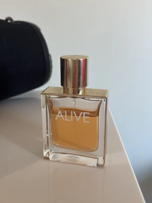 Hugo Boss Alive Eau de Parfum - Alive från Hugo Boss är en elegant parfym i en rektangulär glasflaska med guldigt lock. Doften har en varm bärnstensfärgad vätska och flaskan har en stilren design med texten 'ALIVE' på framsidan. Perfekt för dig som gillar lyxiga och moderna dofter. Använd några gånger💞