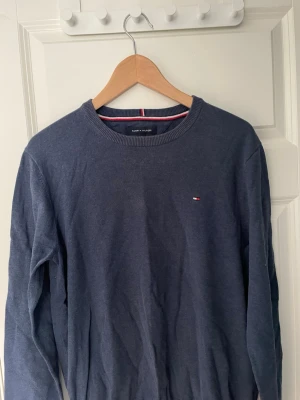 Mörkblå stickad tröja Tommy Hilfiger 100% bomull - Snygg mörkblå stickad tröja från Tommy Hilfiger med rund halsringning och diskret logga broderad på bröstet. Tröjan har ribbade muddar vid hals, ärmslut och nederkant. Perfekt till jeans eller chinos för en clean look. 100% bomull. Storlek M.