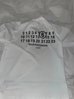 Vit Maison Margiela t-shirt - Säljer en vit t-shirt från Maison Margiela med tryck på både fram- och baksidan. På bröstet finns en liten logga och på ryggen syns det ikoniska numrerade trycket med siffran 6 markerad. T-shirten har rund hals och korta ärmar. Materialet är mjuk bomull.