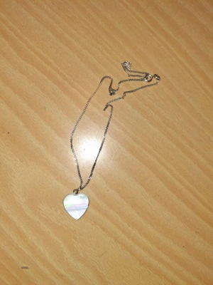 Halsband med hjärthänge i pärlemor - Snyggt halsband med ett hjärtformat hänge i skimrande pärlemor på en tunn silverfärgad kedja. Perfekt accessoar för att lyfta din outfit med en elegant touch. Hjärtat har en mjuk, glansig yta som reflekterar ljuset fint.