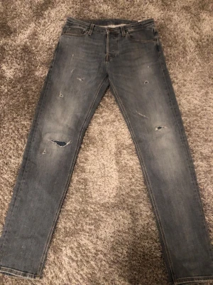 Ljusblåa/gråa slitna jeans från Jack & Jones - Säljer ett par ljusblåa skinny jeans från Jack & Jones med slitningar och hål på benen för en trendig look. Klassisk femficksmodell med dragkedjegylf och läderpatch bak. Perfekta för dig som gillar streetstyle och avslappnad vibe.
