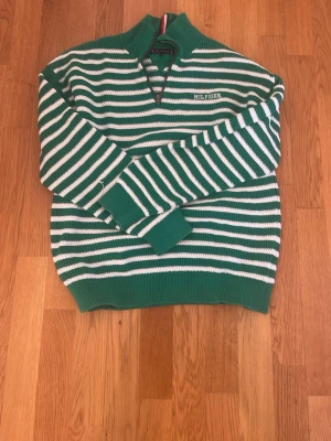 Grönvit randig stickad tröja Tommy Hilfiger - Snygg stickad tröja från Tommy Hilfiger i grönt och vitt med klassiska ränder. Tröjan har halvzip och hög krage, samt logotyp broderad på bröstet. Ribbstickade muddar och nederkant ger en skön passform. Perfekt för dig som gillar stilrena och sportiga plagg. Den har dock lite hål eller lite sönder på sig men de märks inte jag har fotat de som är sönder