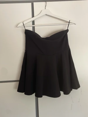 Svart axelbandslös tubtopp från H&M - Snygg svart axelbandslös tubtopp från H&M Divided. Toppen har en bandeauformad överdel och ett utsvängt fall nedtill. Aldrig använd!!