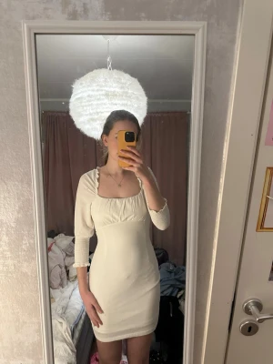 Krämvit bodycon klänning med öppen rygg - Säljer en krämvit bodycon klänning med långa ärmar och fyrkantig urringning. Klänningen har fina volangdetaljer vid axlarna och en öppen rygg med snörning. Materialet är stretchigt och följsamt, perfekt för dig som gillar en figurnära passform.