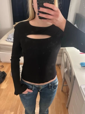 Svart ribbad topp med cut-out - Svart långärmad topp med ribbad struktur och en snygg cut-out detalj vid bröstet. Modellen är figurnära och har lätt utsvängda ärmar. Perfekt festtopp och aldrig använd