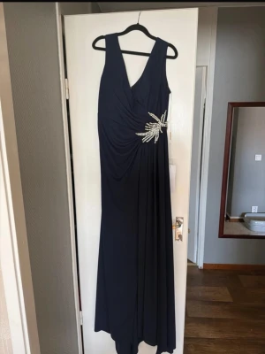 Marinblå långklänning med pärldetalj - Elegant marinblå långklänning med djup v-ringning och breda axelband. Klänningen har en snygg drapering över bysten och midjan, samt en dekorativ pärlbroderad detalj på sidan. Perfekt för speciella tillfällen. Klänningen är helt ny med etikett. Den säljs för att den var för lång. Den passar bra för storlek 44-46 och längd över 160cm. Priset kan diskuteras. 
