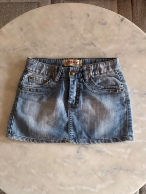 Jeans kjol - Snygg ljusblå jeanskjol med klassisk femficksdesign, nitar och dragkedjor på bakfickorna. Kjolen har bälteshällor och knappstängning framtill. Perfekt för en trendig och avslappnad look.