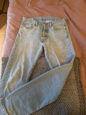 Levi's 501 ljusblå raka jeans - Klassiska Levi's 501 jeans i ljusblå denim med raka ben och femficksmodell. Jeansen har knappgylf och den ikoniska läderpatchen bak i midjan. Perfekta för en avslappnad och tidlös stil. Byxorna har mindre slitningar men är fortfarande fräscha, storlek 30 32
