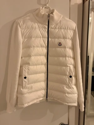 Vit cardigan från Moncler - Snygg vit dunjacka från Moncler med stickade ärmar och quiltad front. Jackan har två fickor med knappar, hög krage och dragkedja framtill. Moncler-logga på bröstet och broderad detalj på ärmen. Perfekt för kalla vinterdagar. Storlek M men passar S också. 
