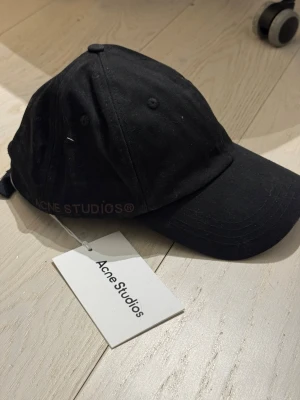 Svart keps från Acne Studios - Snygg svart keps från Acne Studios med broderad logga på sidan. Klassisk böjd skärm och justerbar rem baktill. Tillverkad i bomull för en bekväm och stilren look. Perfekt accessoar för att lyfta din outfit.