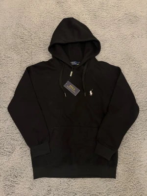 Svart hoodie från Polo Ralph Lauren - Klassisk svart hoodie från Polo Ralph Lauren med broderad logga på bröstet. Tröjan är helt ny, aldrig använd utan bara testad. Tröjan är i storlek M men passar bättre Xs/S (160-174cm). Priset är ej hugget i sten så kom gärna med bud!