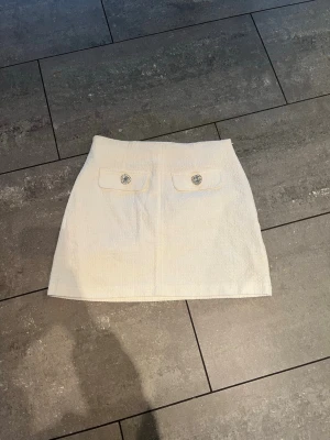 Vit kortkjol med fickdetaljer - Stilren vit kortkjol med två fickor framtill, dekorerade med glittriga knappar. Kjolen har en rak passform och är tillverkad i ett strukturerat tyg som ger en exklusiv känsla. Perfekt att matcha med en snygg topp eller stickad tröja.