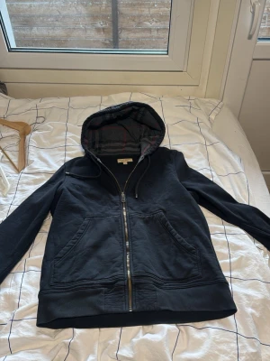 Svart hoodie från Burberry med dragkedja - Säljer en svart hoodie från Burberry med klassisk dragkedja och två stora fickor framtill. Huva med ikoniskt rutmönster i grått och rött på insidan. Diskret Burberry-logga på bröstet. Perfekt för dig som vill ha en stilren och exklusiv look.