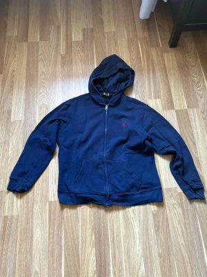 Marinblå hoodie från Polo Ralph Lauren - Säljer en marinblå hoodie från Polo Ralph Lauren med dragkedja och huva. Klassisk design med röd logobrodyr på bröstet och stora fickor framtill. Perfekt för dig som gillar stilrena och bekväma plagg. Materialet är mjukt och skönt, passar till chill dagar.