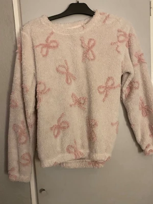 Ljusrosa fluffig loungewear-tröja - Mysig och fluffig loungewear-tröja i ljusrosa med söta rosett-mönster i en mörkare rosa nyans. Tröjan har rund hals och långa ärmar, perfekt för att chilla hemma. Materialet är mjukt och ger en skön känsla mot huden.