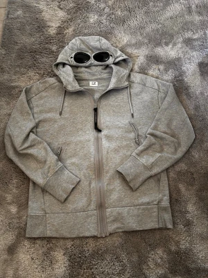 Grå hoodie med glasögon från C.P. Company - Säljer en grå hoodie från C.P. Company med dragkedja framtill och ikoniska inbyggda glasögon i huvan. Hoodien har två fickor med dragkedja och ribbade muddar. Tillverkad i mjukt bomullsmaterial och har en modern, avslappnad passform.