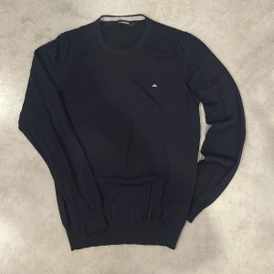 Mörkblå stickad tröja från J.Lindeberg - Snygg mörkblå stickad tröja från J.Lindeberg med rund halsringning och diskret logga broderad på bröstet. Tröjan har långa ärmar och ribbade muddar vid ärmslut och nederkant. Perfekt för lager-på-lager och stilren look.