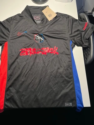 Ny FC Barcelona x Nike x Travis Scott fotbollströja svart/röd/blå – med tags – aldrig använd - Snygg och unik fotbollströja från FC Barcelona i samarbete med Nike och Travis Scott. Tröjan är helt ny och oanvänd med original tags kvar. Modern svart design med röda och blå detaljer samt Cactus Jack-tryck framtill. Perfekt för både match, träning eller streetwear.  • Skick: Aldrig använd (helt ny) • Tags kvar • Inget tryck på baksidan • Lätt och bekvämt material • Perfekt present eller samlarplagg  fotbollströja, Barcelona tröja, Nike jersey, Travis Scott merch, svart fotbollströja