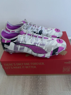 Puma vita/lila fotbollsskor mönstrade - Säljer ett par riktigt snygga fotbollsskor från Puma i vitt med lila, svart och rosa grafiskt mönster. Skorna har klassisk snörning, låg profil och är tillverkade i ett lätt syntetmaterial för bästa känsla på planen. Perfekta för dig som vill sticka ut på matchen!