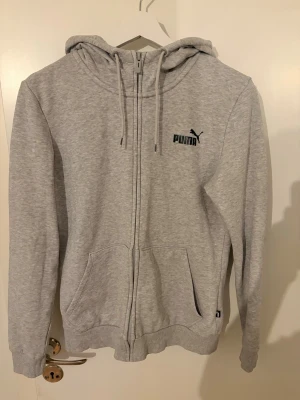 Grå zip hoodie från Puma - Snygg grå hoodie från Puma med dragkedja framtill och två fickor. Klassisk logga på bröstet och justerbar huva med snören. Tillverkad i mjukt sweatshirtmaterial som är perfekt för chill dagar. Ribbade muddar vid ärmslut och nederkant.