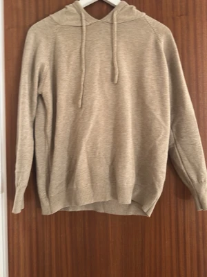 Beige merinoull hoodie - En stilren beige hoodie med huva. Hoodien har långa ärmar och är tillverkad i ett mjukt, stickat material som känns skönt mot huden. Perfekt för en avslappnad look.