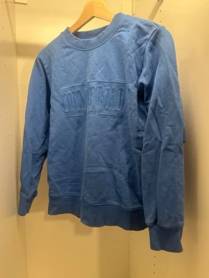 Blå sweatshirt från Country Road - Blå sweatshirt med broderad text 'Country Road 1974' på bröstet. Tröjan har rund hals, ribbade muddar vid ärmslut och nederkant samt en avslappnad passform. Perfekt för en chill och stilren look.