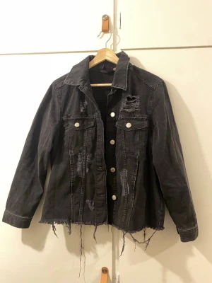 Svart/grå jeansjacka - Säljer en svart jeansjacka med slitna detaljer och rå kant nertill. Jackan har två bröstfickor med lock och silverfärgade knappar framtill. Oversized passform och cool vintagekänsla, perfekt för dig som gillar streetstyle. Inga tecken på användning! 