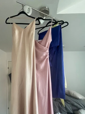 Champagnefärgad långklänning i satin - Elegant långklänning i champagnefärgad satin med smala axelband och v-ringad halsringning. Klänningen har en figurnära passform och ett stilrent, minimalistiskt uttryck som ger en lyxig känsla.