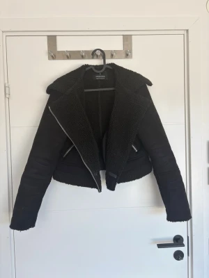 Svart pälsjacka från Stradivarius - Snygg svart pälsjacka från Stradivarius med bred krage och dragkedja. Jackan har fuskpäls på insidan och detaljer vid ärmslut och nederkant. Kortare modell med två fickor framtill och cool bikerstil. Perfekt för kalla dagar. Mocka uttill