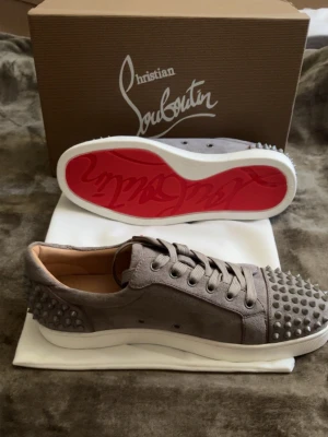 Grå Christian Louboutin sneakers - Snygga grå sneakers från Christian Louboutin i mocka med ikoniska nitar på tå och häl. Skorna har klassisk röd sula, vita kanter och grå snörning. Insidan är i beige läder för extra komfort. Perfekta för dig som vill sticka ut med exklusiv stil.