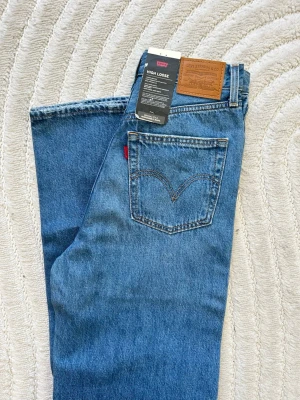 Levi's High Loose blå jeans - Säljer ett par klassiska blå jeans från Levi's i modellen High Loose. Jeansen har hög midja, lös passform och femficksdesign. Tillverkade i slitstark denim med ikonisk läderpatch bak och röd Levi's-tagg på bakfickan. Helt nya och aldrig använda! Prislappen sitter fortfarande på! I storlek 26x33😍