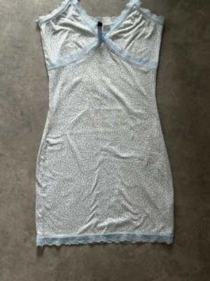 Blommigt nattlinne med spets från H&M - Sött nattlinne från H&M Divided i storlek S. Ljusblå spetsdetaljer vid halsringning och nederkant, samt ett litet blått rosett framtill. Tunt material med småblommigt mönster i blått och grönt på vit botten. Perfekt för mysiga kvällar hemma.