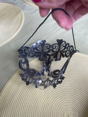 Svart spetsmask med kristaller - Elegant maskeradmask i transparent plast dekorerad med svart spets och små kristaller. Masken har ett snörband för att fästa runt huvudet och detaljerade mönster som ger en mystisk look. Perfekt för cosplay eller maskerad.snörenaband kan man byta gummiband. Då sitter den bättre 