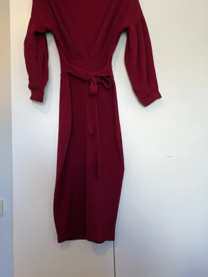 Vinröd långklänning med knytband - Vinröd långklänning med v-ringad omlott-topp, långa ärmar och knytband i midjan. Klänningen har en elegant och enkel design med markerad midja och mjukt, strukturerat tyg som ger en snygg siluett.