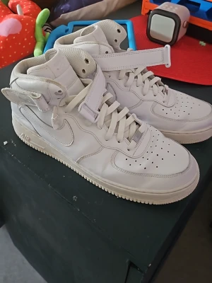 Vita Nike Air Force 1 High.. size 46 - Klassiska vita Nike Air Force 1 High sneakers i läder med högt skaft, perforerad tåbox och kardborreband runt ankeln. Swoosh-logga på sidan och tjock vit sula. Perfekta för en clean och sportig look.