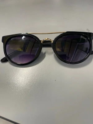 Svarta solglasögon från Dior - Snygga svarta solglasögon från Dior med rundade bågar och mörka glas. Bågarna har en cool gulddetalj vid näsryggen och en tunn guldfärgad metallbåge upptill. Perfekta för dig som vill ha en trendig och exklusiv look.
