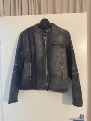 Svart skinnjacka från Eytys (Damon) - Säljer en svart skinnjacka från Eytys med coolt sliten finish och vita kontrastsömmar. Jackan har ståkrage med tryckknapp, dragkedja framtill, tre fickor med dragkedja och broderad Eytys-logga på ryggen. Snygga detaljer på ärmarna och schysst bikerstil.