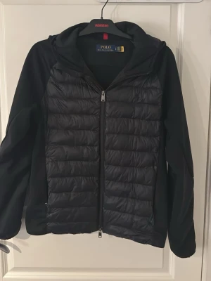 Svart pufferjacka från Polo Ralph Lauren - Säljer en svart cardigan från Polo Ralph Lauren med quiltad front och släta ärmar.  Materialet är en kombination av syntet och dun, perfekt för kalla vinterdagar. Litet skrapsår på armbågen men inget man riktigt märker av. Pris kan diskuteras.