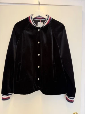 Svart bomberjacka från Tommy Hilfiger - Snygg svart bomberjacka i sammet från Tommy Hilfiger med vita, röda och blå ränder på krage, ärmslut och rygg. Jackan har tryckknappar framtill och två fickor. Perfekt för dig som vill ha en sportig och trendig look.