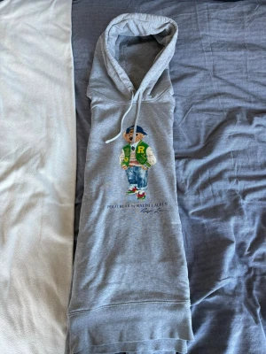 Grå hoodie Polo Bear Ralph Lauren - Snygg grå hoodie från Ralph Lauren med Polo Bear-tryck framtill. Klassisk huva med dragsko och ribbade muddar. Perfekt för dig som gillar streetwear och vill ha en ikonisk look. Mjuk bomullsblandning och avslappnad passform.