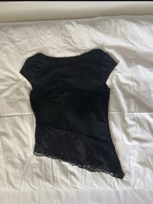 Svart asymmetrisk topp med spets - Cool svart topp med korta ärmar och asymmetrisk nederkant. Den har snygga spetsdetaljer både upptill och längs nederkanten, vilket ger en unik look. Perfekt för dig som gillar att sticka ut med detaljer och stil.