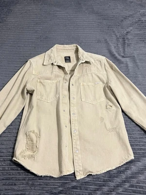 Beige overshirt från Zara med slitningar - Stilren beige overshirt från Zara med två bröstfickor och slitna detaljer för en trendig look. Skjortan har långa ärmar, knappar framtill och är tillverkad i ett mjukt bomullsmaterial. Perfekt för dig som gillar en avslappnad och edgy stil.
