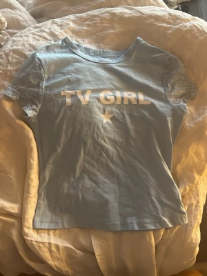 Blå TV Girl t-shirt med spetsärmar - Ljusblå t-shirt med texten 'TV GIRL' och en stjärna framtill. T-shirten har korta ärmar i spets som ger en unik touch. Ingen storlek men skulle säga S-M
