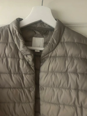 Beige dunjacka med knappar - Säljer en beige dunjacka med rund hals och knäppning framtill. Jackan har ett quiltat mönster och är lätt vadderad, perfekt för kalla vinterdagar. Materialet är syntet och dun, och modellen är klassisk med långa ärmar.